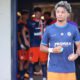 Young male soccer player in a blue jersey holding a ball, standing near a tunnel with teammates in orange shorts in the background.