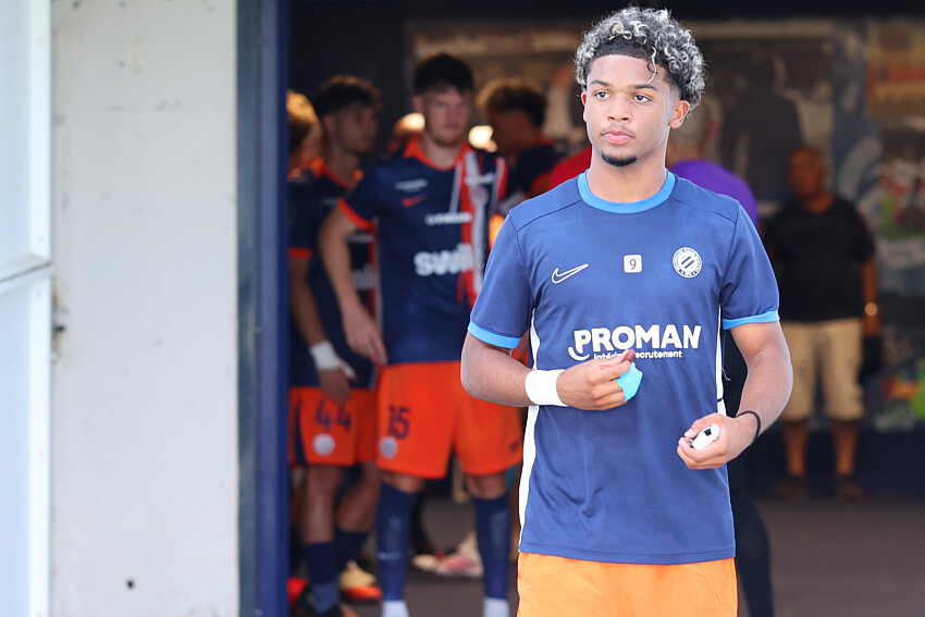 Mégnan | AllezPaillade.com Young male soccer player in a blue jersey holding a ball, standing near a tunnel with teammates in orange shorts in the background.