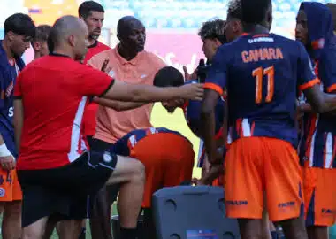 Soccer team huddles around coach during a break on the field, players in orange and navy kits listening intently.