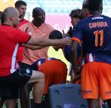 Soccer team huddles around coach during a break on the field, players in orange and navy kits listening intently.