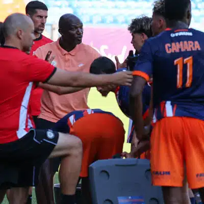 Soccer team huddles around coach during a break on the field, players in orange and navy kits listening intently.