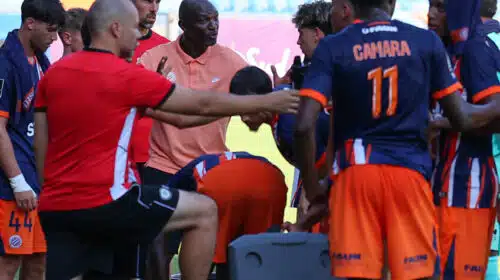 Soccer team huddles around coach during a break on the field, players in orange and navy kits listening intently.