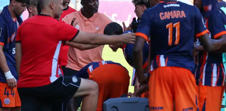 Camara | AllezPaillade.com Soccer team huddles around coach during a break on the field, players in orange and navy kits listening intently.