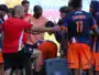 Soccer team huddles around coach during a break on the field, players in orange and navy kits listening intently.