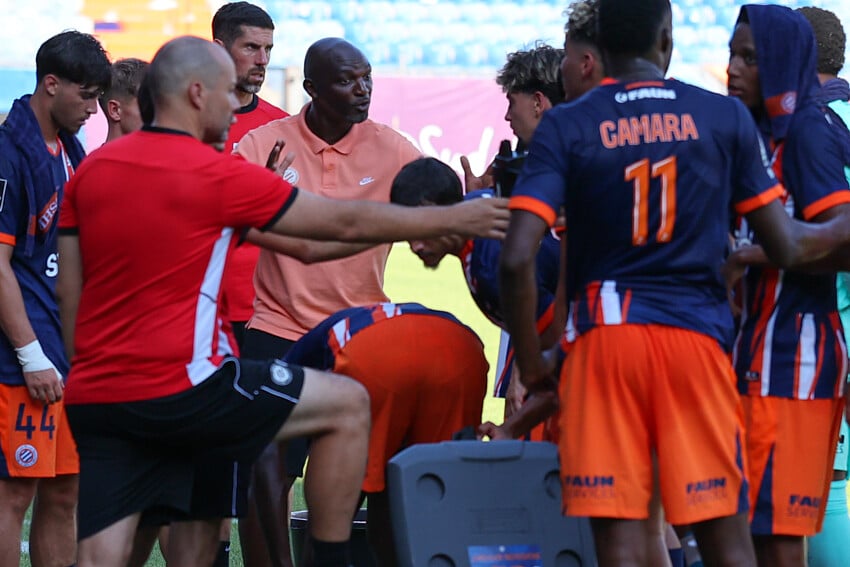 Camara | AllezPaillade.com Soccer team huddles around coach during a break on the field, players in orange and navy kits listening intently.
