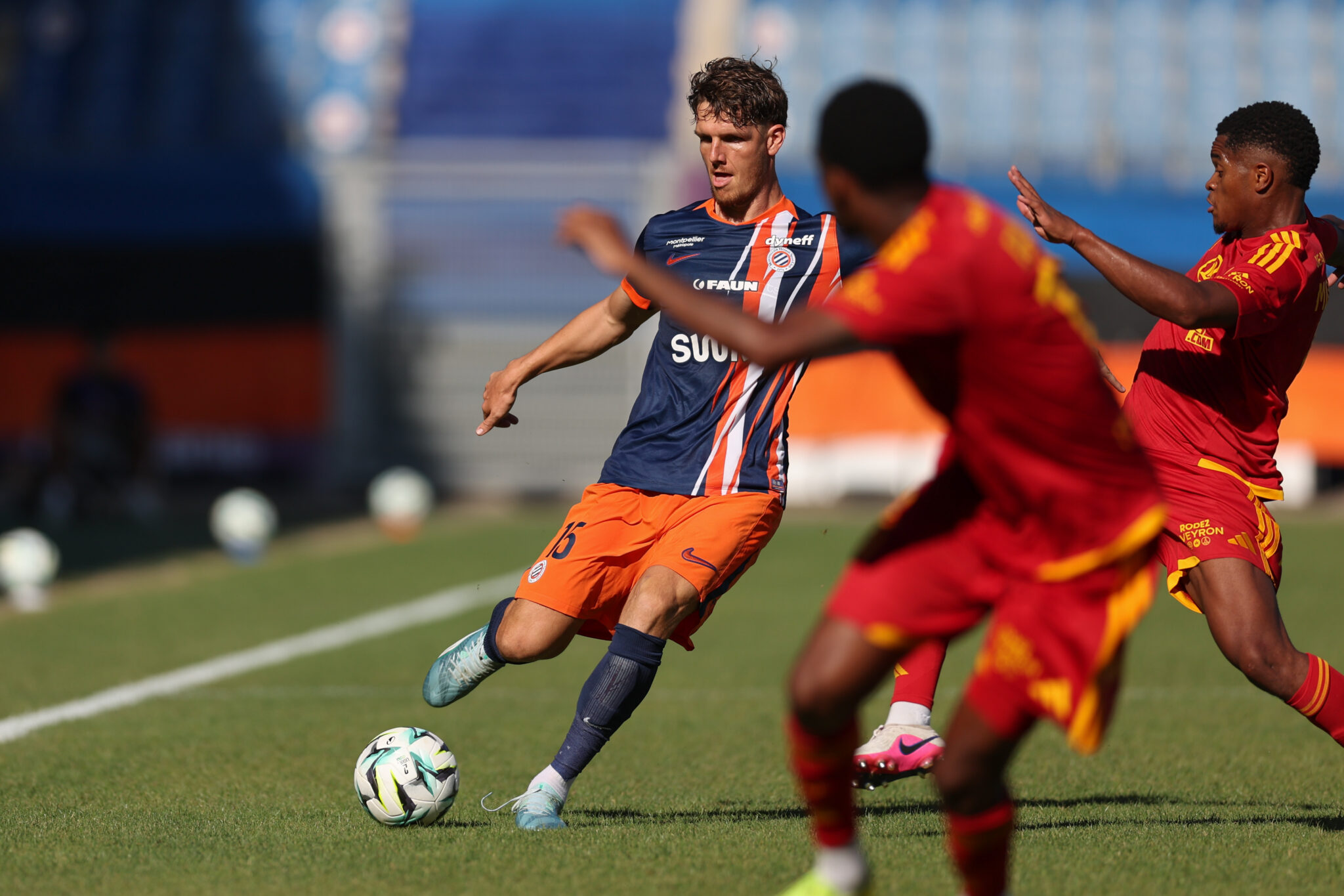 Julien Laporte Montpellier Herault SC v Rodez Aveyron Football - Friendly Match 01082026 | AllezPaillade.com Soccer players in blue/orange and red uniforms chase a ball on a grassy pitch during a match, with one player in blue controlling the ball near the foreground.