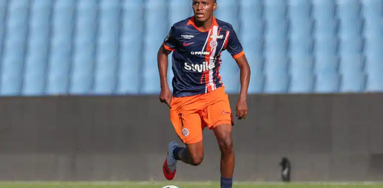 Yanis Issoufou Montpellier Herault SC v Rodez Aveyron Football - Friendly Match 01082026 | AllezPaillade.com Soccer player in navy and orange kit dribbling a ball on a green field with stadium seats in the background