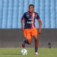 Soccer player in navy and orange uniform dribbles a ball on a green field with blue stadium seats behind him.