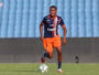Soccer player in navy and orange uniform dribbles a ball on a green field with blue stadium seats behind him.