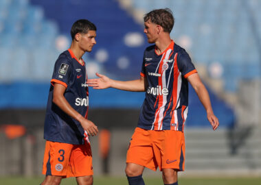 Two soccer players in navy and orange kits greet each other with a handshake on the field during a match.