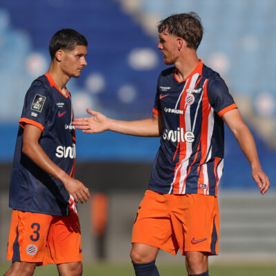 Two soccer players in navy and orange kits greet each other with a handshake on the field during a match.