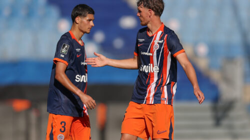 Two soccer players in navy and orange kits greet each other with a handshake on the field during a match.