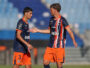 Two soccer players in navy and orange kits greet each other with a handshake on the field during a match.