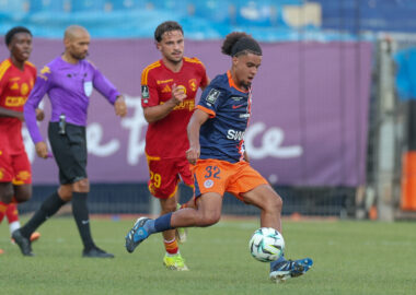 Soccer match action with a blue-jersey player dribbling the ball as red-jersey opponents chase behind on a green field.