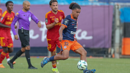 Soccer match action with a blue-jersey player dribbling the ball as red-jersey opponents chase behind on a green field.