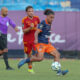 Soccer match action with a blue-jersey player dribbling the ball as red-jersey opponents chase behind on a green field.