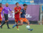 Soccer match action with a blue-jersey player dribbling the ball as red-jersey opponents chase behind on a green field.