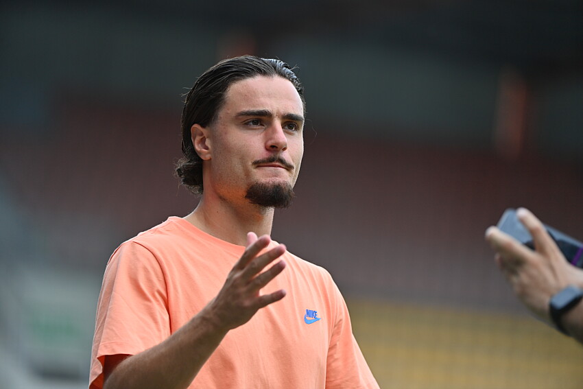 Pays | AllezPaillade.com Man with dark hair and a goatee wearing a peach Nike T-shirt gestures while being filmed by a person with a phone.