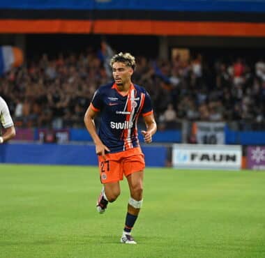 Soccer player in navy orange kit dribbles on a green field as a grey-clad opponent closes in; crowded stadium in the background.