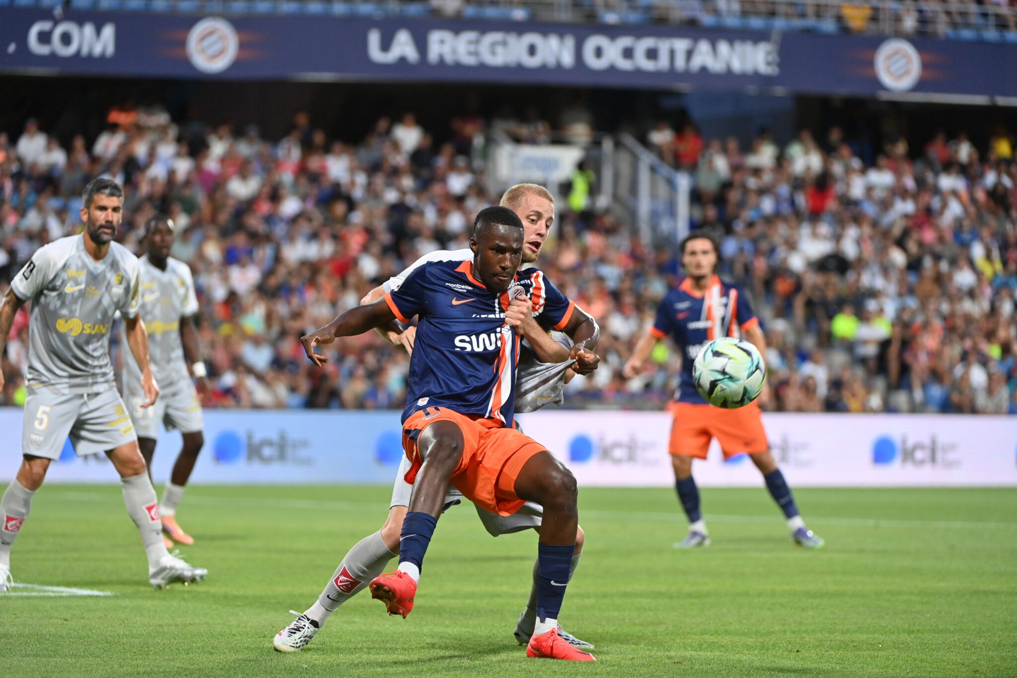 Mamadou CAMARA Photo by Icon Sport - Photo by Icon Sport | AllezPaillade.com Soccer match in progress with a player in blue and orange sprinting for the ball while a defender in white pursues, in a packed stadium.