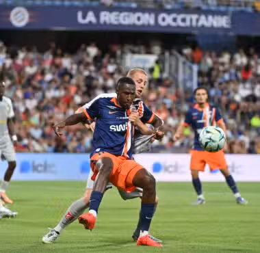 Soccer match in progress with a player in blue and orange sprinting for the ball while a defender in white pursues, in a packed stadium.