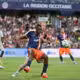 Soccer match in progress with a player in blue and orange sprinting for the ball while a defender in white pursues, in a packed stadium.