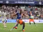 Soccer match in progress with a player in blue and orange sprinting for the ball while a defender in white pursues, in a packed stadium.