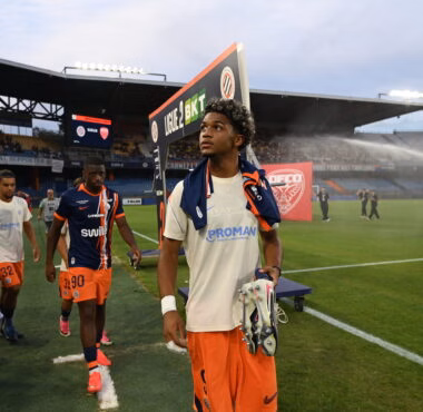 Soccer players walk off the field near the sideline with a stadium crowd in the background.