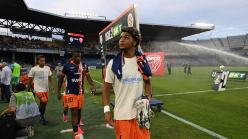 Soccer players walk off the field near the sideline with a stadium crowd in the background.