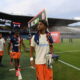 Soccer players walk off the field near the sideline with a stadium crowd in the background.