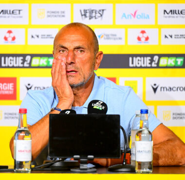 Man at a press conference, wearing a light blue polo, resting his head on his hand in front of a yellow sponsor wall and microphones.