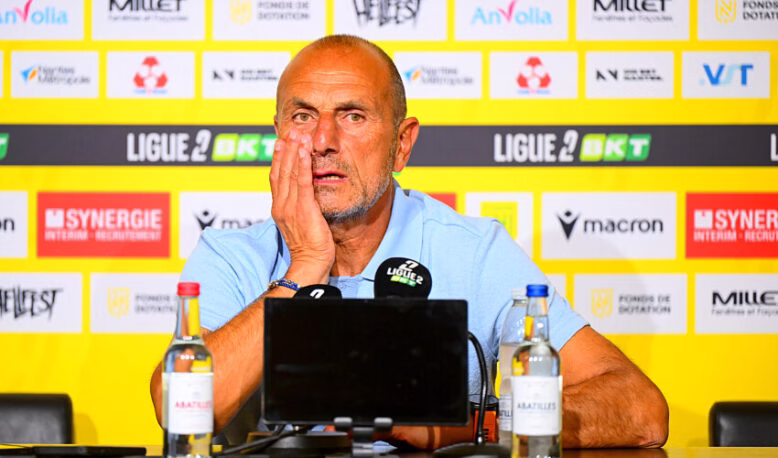 Man at a press conference, wearing a light blue polo, resting his head on his hand in front of a yellow sponsor wall and microphones.