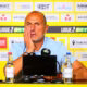 Man at a press conference, wearing a light blue polo, resting his head on his hand in front of a yellow sponsor wall and microphones.