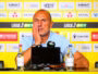 Man at a press conference, wearing a light blue polo, resting his head on his hand in front of a yellow sponsor wall and microphones.