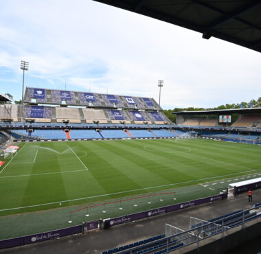 Empty soccer stadium with a green pitch, blue seats, and banners along the stands under a clear sky.