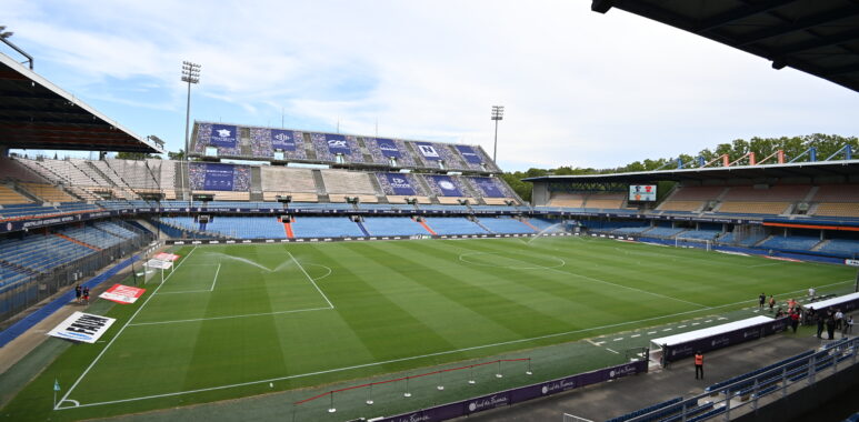 Empty soccer stadium with a green pitch, blue seats, and banners along the stands under a clear sky.