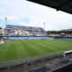 Empty soccer stadium with a green pitch, blue seats, and banners along the stands under a clear sky.