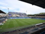 Empty soccer stadium with a green pitch, blue seats, and banners along the stands under a clear sky.
