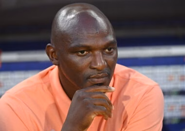 Close-up of a man in a peach shirt resting his chin on his hand, looking pensive.