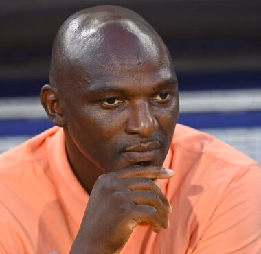Close-up of a man in a peach shirt resting his chin on his hand, looking pensive.
