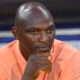 Close-up of a man in a peach shirt resting his chin on his hand, looking pensive.