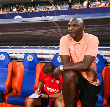 Man in peach polo sits on a stadium bench with orange and blue seats, watching a game as a teammate writes notes nearby.
