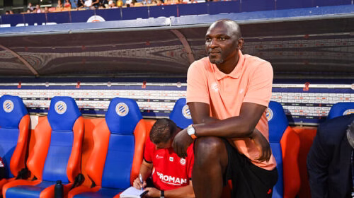 Man in peach polo sits on a stadium bench with orange and blue seats, watching a game as a teammate writes notes nearby.