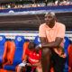 Man in peach polo sits on a stadium bench with orange and blue seats, watching a game as a teammate writes notes nearby.