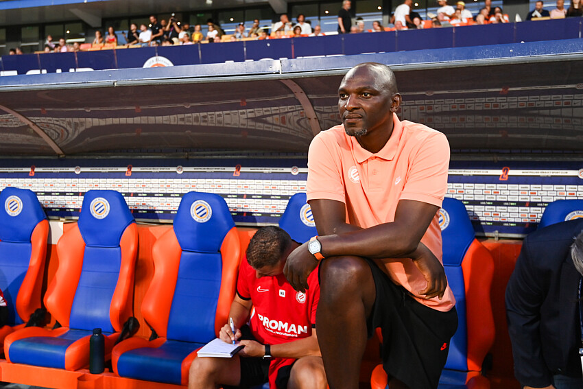 Man in peach polo sits on a stadium bench with orange and blue seats, watching a game as a teammate writes notes nearby.