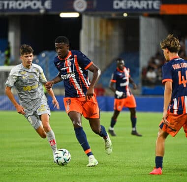 Soccer match: a gray-clad player dribbles the ball while a blue-and-orange team pursues him on the field near the goal area.