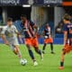 Soccer match: a gray-clad player dribbles the ball while a blue-and-orange team pursues him on the field near the goal area.
