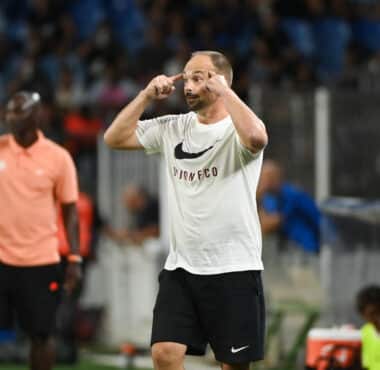Soccer player in a white Nike shirt and black shorts celebrates on the field, fingers to temples in a gesture of focus or celebration.
