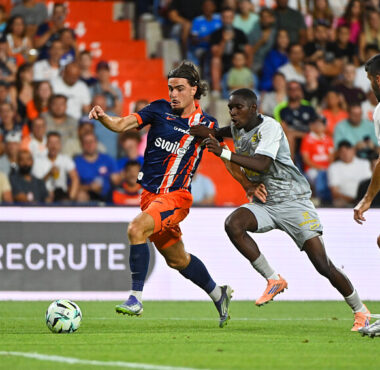 Soccer player in blue-and-orange striped jersey dribbles the ball as two gray-clad defenders close in on a green field with a cheering crowd behind.