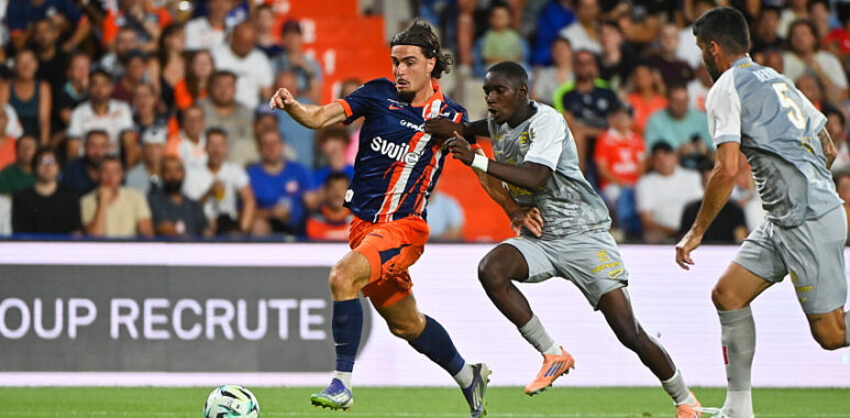 Pays | AllezPaillade.com Soccer player in blue-and-orange striped jersey dribbles the ball as two gray-clad defenders close in on a green field with a cheering crowd behind.